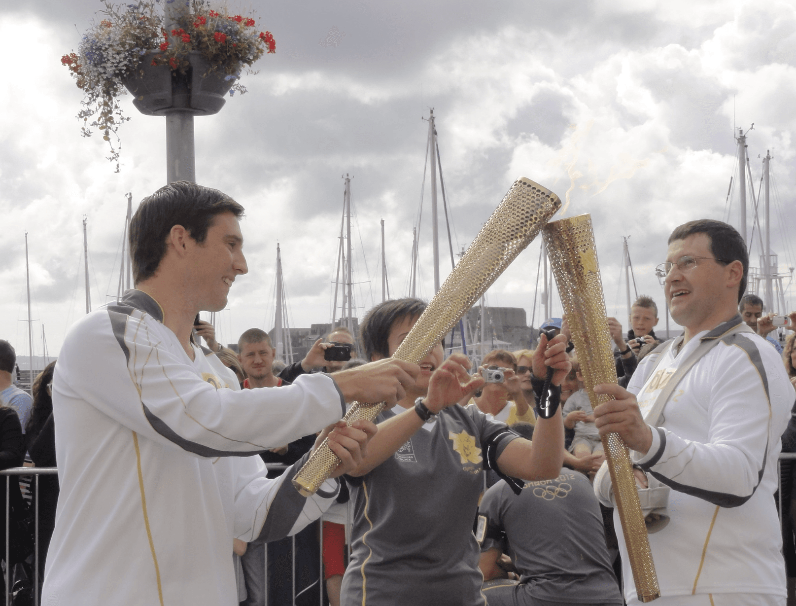Image of two men in white track suits passing the Olympic fire from golden torches in front of a crowd and cloudy sky.